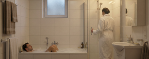 In einem Badezimmer liegt ein Mann in der Badewanne und eine Frau ist im Begriff eine Dusche zu nutzen